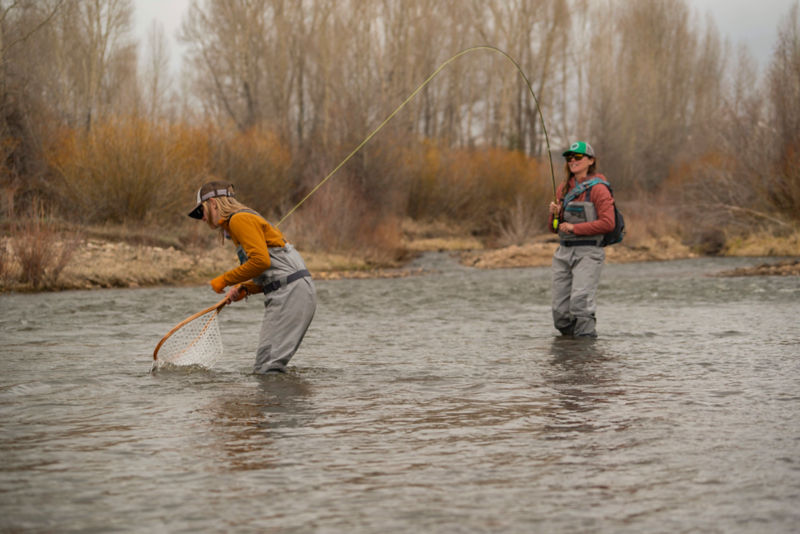 An angler bends down to net a fellow angler's catch from knee-deep in a river.