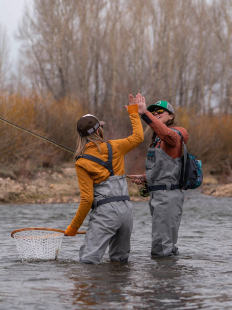 Two women high fiving in a rive while wearing waders and holding fly rods