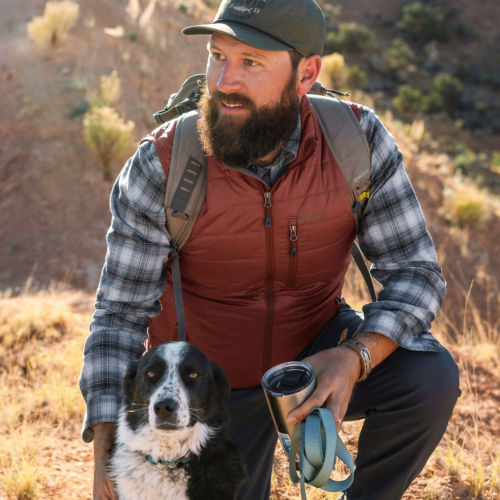 A hiker in a puffy vest kneels next to his black and white dog.