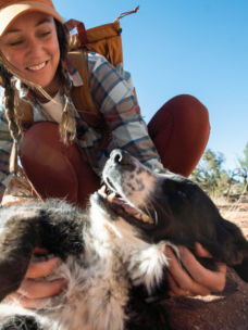 A smiling hiker squats down to ruffle her dog's fur.