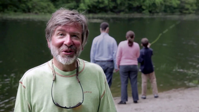 Tom Rosenbauer speaks to the camera. Behind him, parents watch their child cast into a pond.