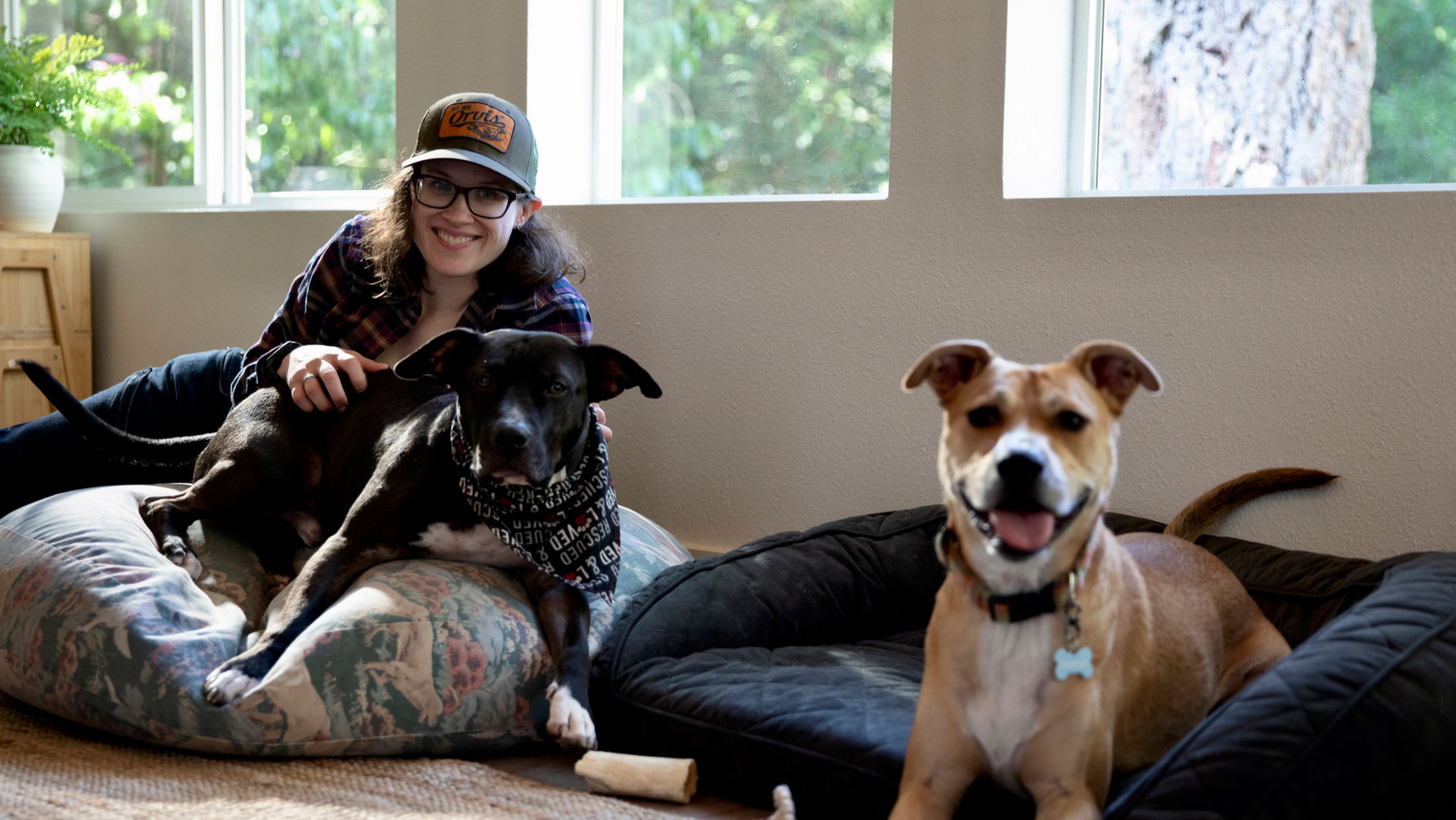 A woman wearing an Orvis baseball cap lounges with her two dogs on their dog beds