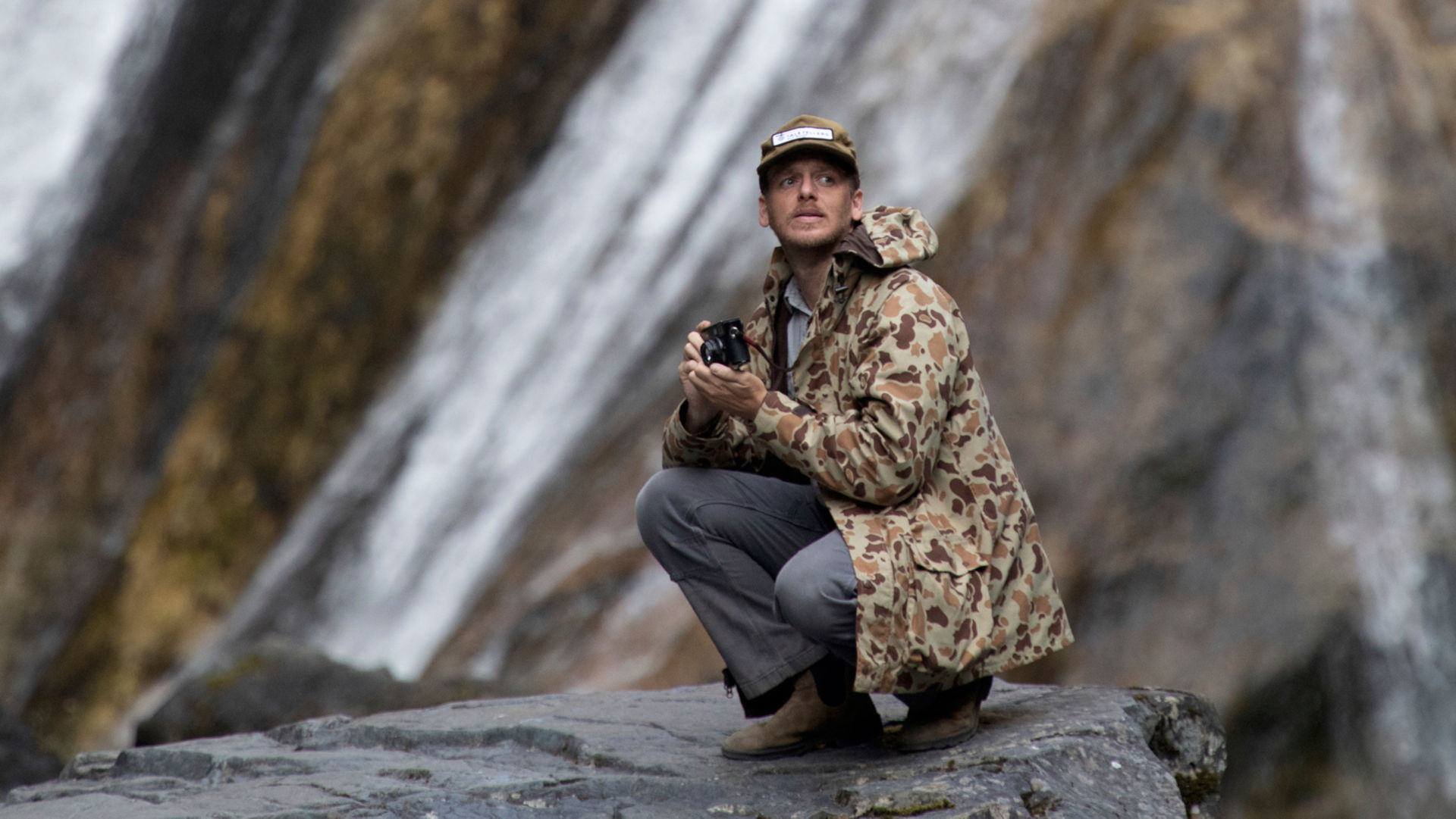 A man in a '71 Camo Jacket squats on a rock in the mountains with a camera in his hand