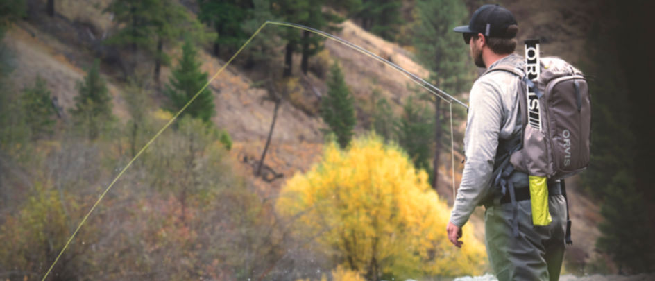 A man wearing an Orvis backpack fly-fishing in a river.