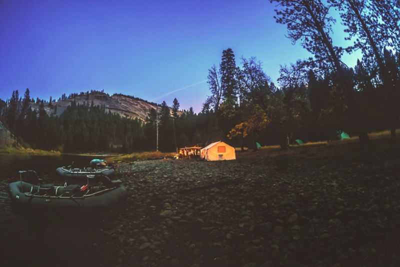 Evening on a riverbank with a lit up tent.