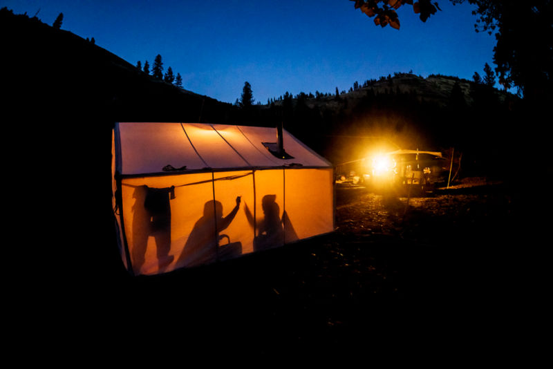 Two people's shadows darken the bright sides of a lit up tent