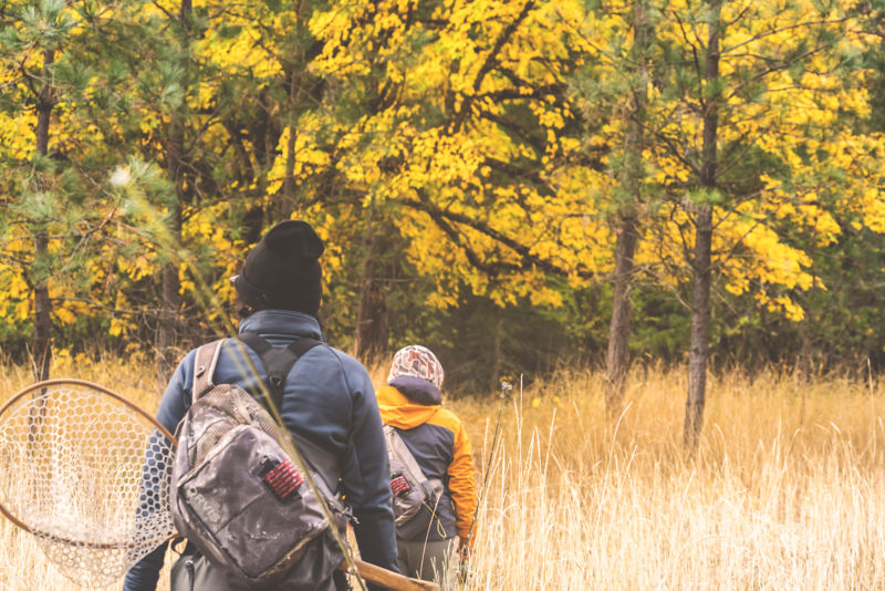 Anglers wearing backpacks head to the water with fly rods in hand.