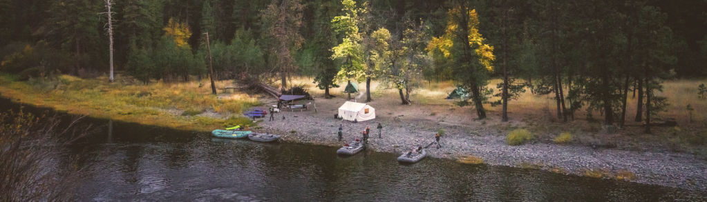 A drone view of several tents set up next to a river with several boats on the riverbank.
