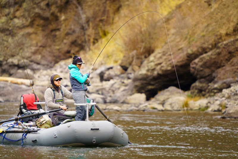 An angler standing and fishing from a raft while her guide steers.