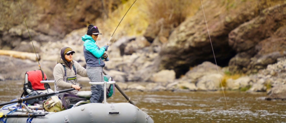An angler standing and fishing from a raft while her guide steers.