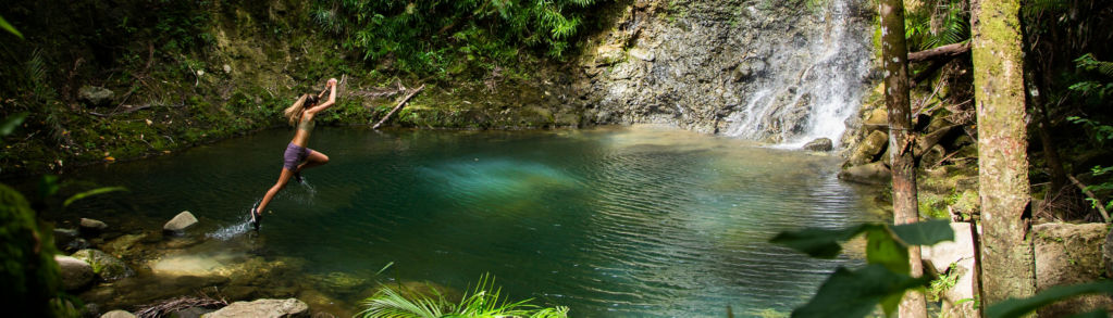 A woman jumps off rocks surrounding a green pool of water beneath a waterfall.