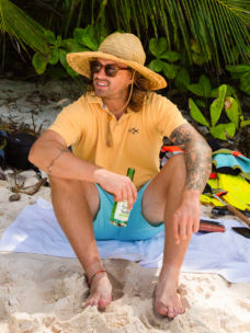 A man in a butter yellow polo shirt sits on a towel on a sandy beach.