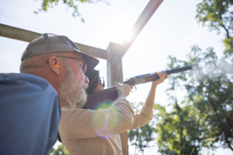 An instructor looks carefully as a student shoots a shotgun.