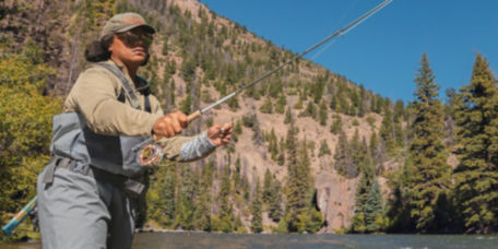 An angler casts her rod from knee-deep in a river surrounded by mountains.