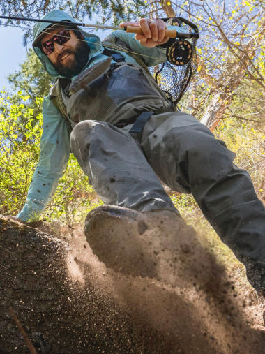 An angler in waders climbs down a dirt bank spraying dirt.