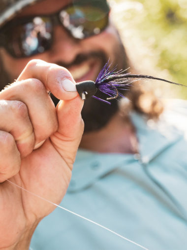 A smiling angler holds up a purple and black fly.