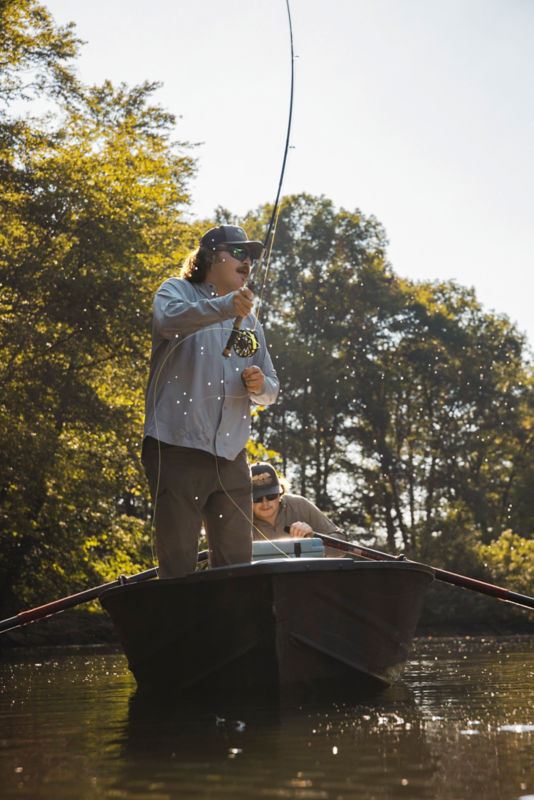 An angler in a ball cap stands in the front of a rowboat.