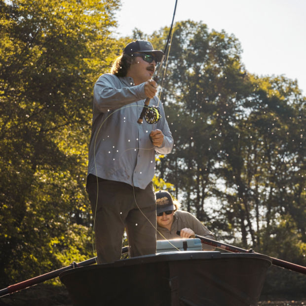 An angler in a ball cap stands in the front of a rowboat.