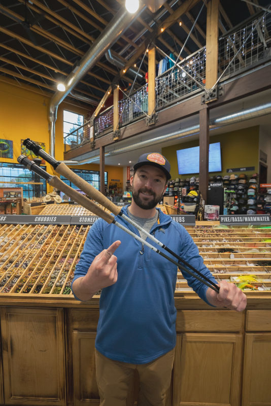 A man stands in front of a fly display holding two fly rods in one hand and pointing to them with his other hand.
