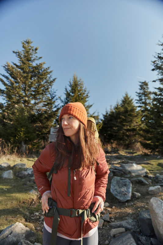 A woman wearing a salmon-colored mid-weight jacket and winter hat while hiking outside