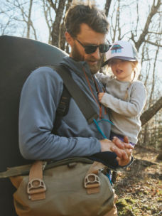 A bearded man carries his child while hiking