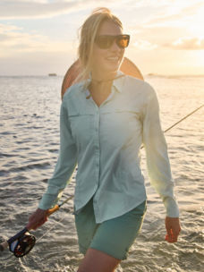A woman wades through the ocean carrying a fly rod.
