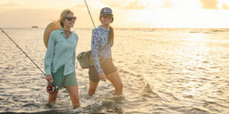 Two women wading through shallow water carrying fishing rods.