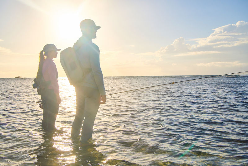 Two anglers stand in the ocean, silhouetted by bright sunlight.
