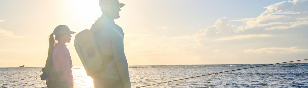 Two anglers stand in the ocean, silhouetted by bright sunlight.