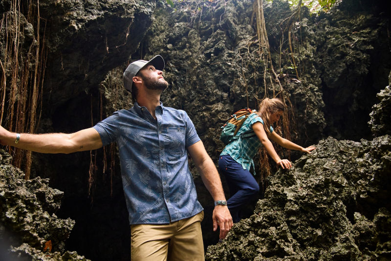 A man wearing UPF clothing stands in a cavern while a woman climbs the rocks.