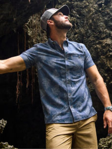 A hiker in a patterned button-down shirt pauses to look up while leaning against a cliff.