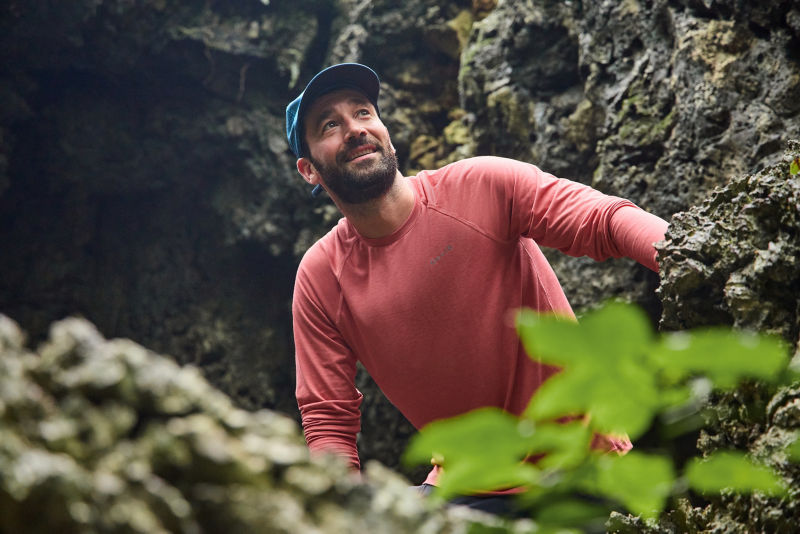 A man wearing a red DriCast crew neck shirt looks up towards the sunlight from a rocky cavern.