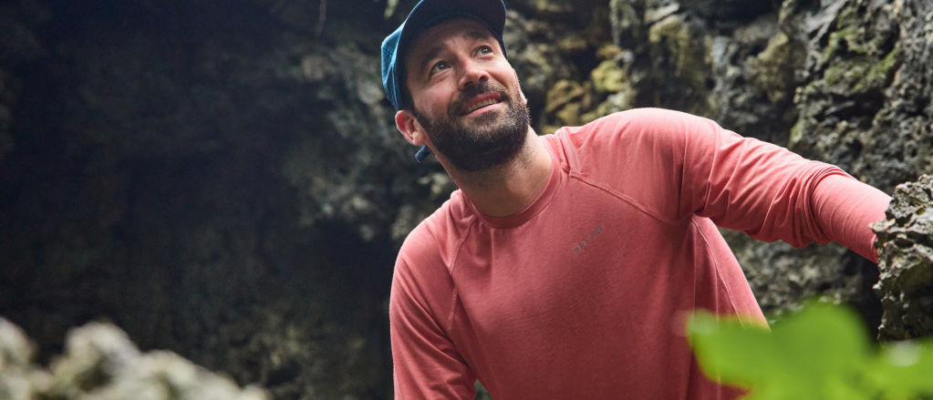 A man wearing a red DriCast crew neck shirt looks up towards the sunlight from a rocky cavern.