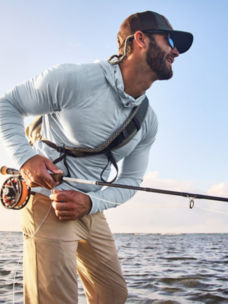 A smiling angler with a sling pack fights a fish.