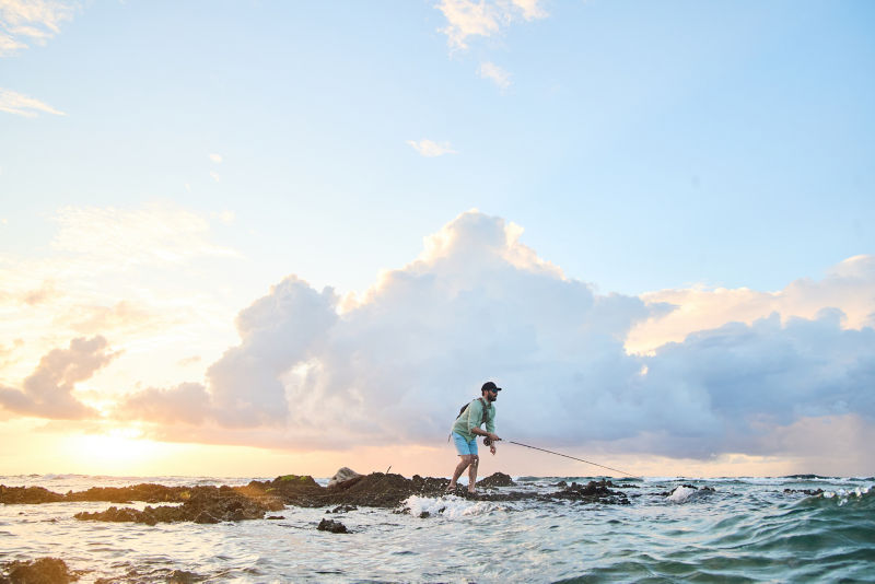 An angler casts into the surf from a rock outcropping under a dramatic sky.