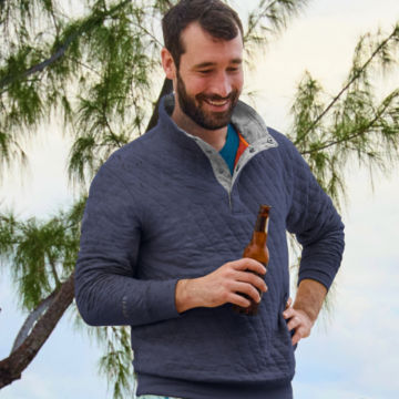 A man wearing an outdoor quilted snap sweatshirt drinks a drink on the beach.