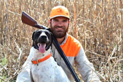 Hunter sitting in a field, wearing orange, with his gun and dog.