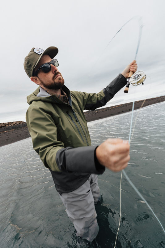 An angler pulls back his rod from knee-deep in a stream on a gray day.