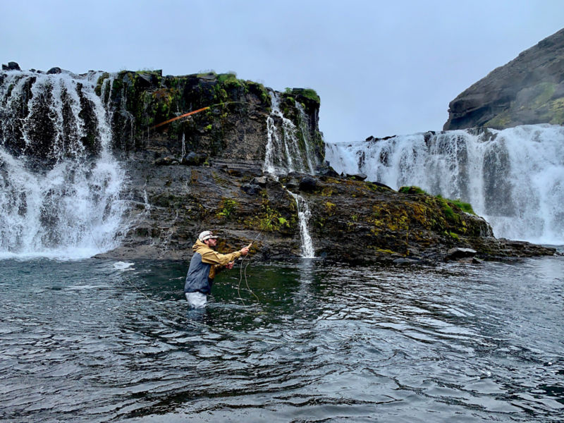A man fly fishing in a rocky river with waterfalls in the background