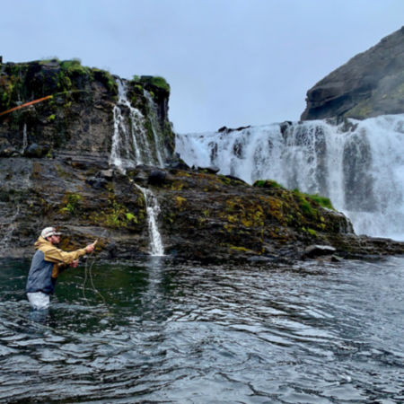 A man fly fishing in a rocky river with waterfalls in the background
