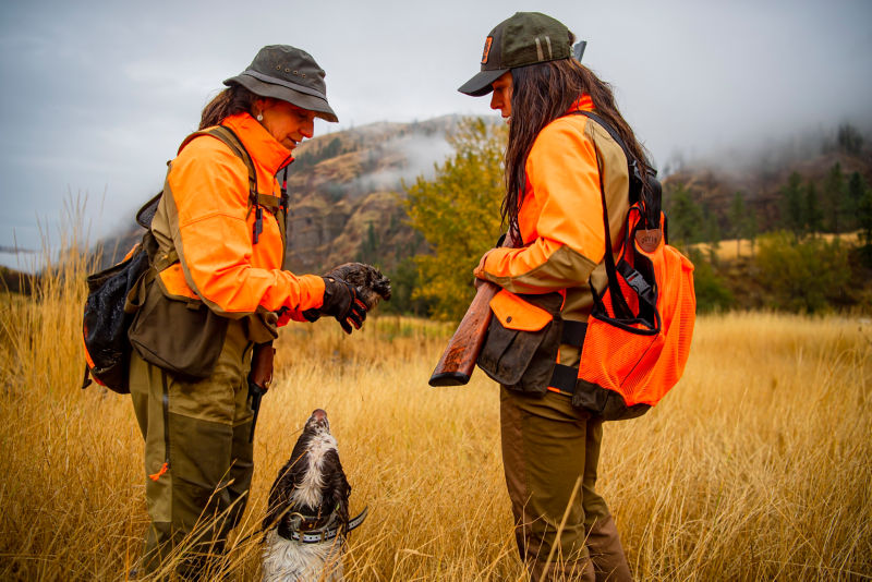 Two women in blaze orange pause with a dog in a golden field.