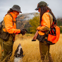 Two women in blaze orange pause with a dog in a golden field.
