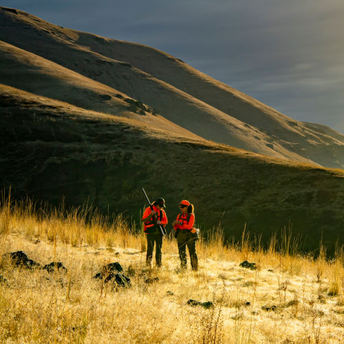 Two hunters standing in a field with the mountains behind them