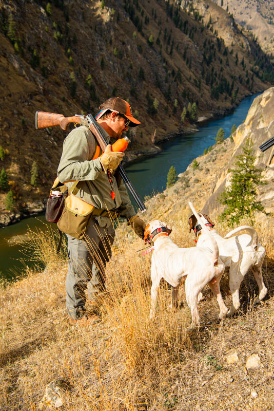 A hunter and their two dogs on a steep hill with the river below.