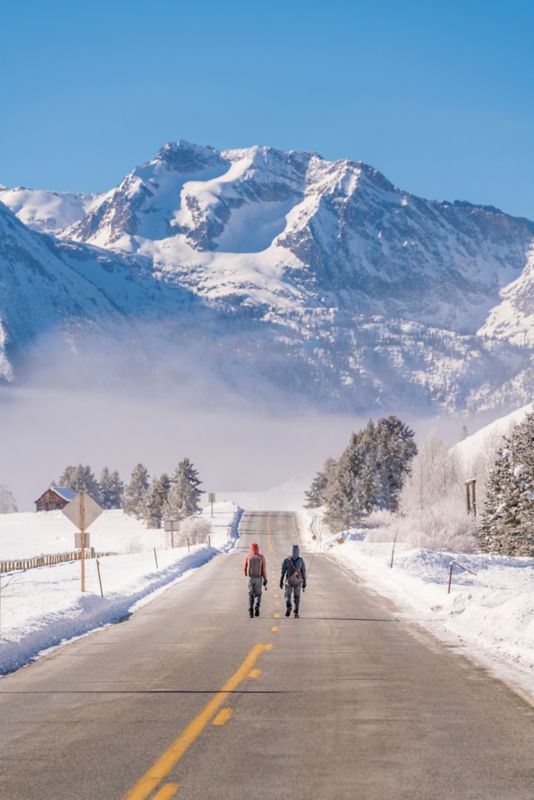 A man and a woman dressed in fishing gear walking on a road toward mountains in the winter.
