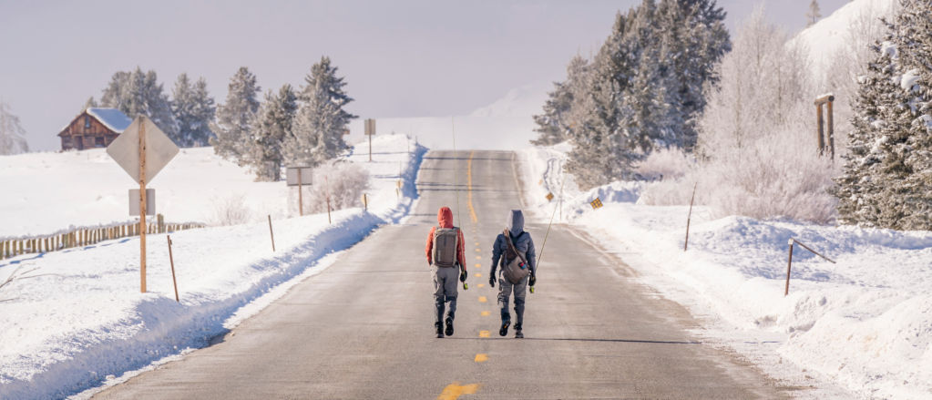 A man and a woman dressed in fishing gear walking on a road toward mountains in the winter