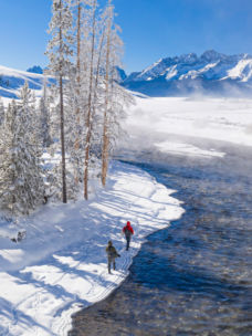 Two anglers walking along a river on a snowy, misty morning.