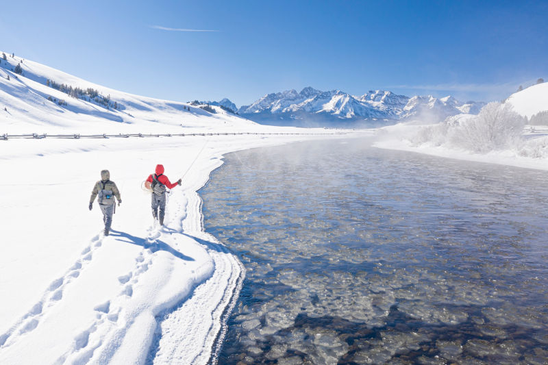Two anglers trek through the snow on the side of a stream.