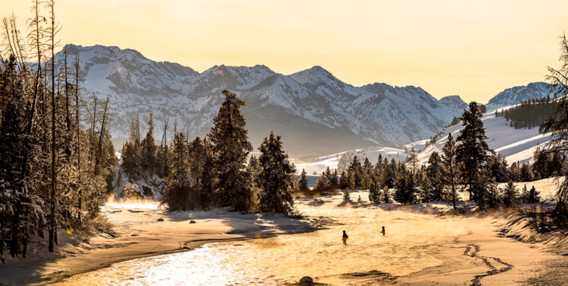 An aerial view of two people fishing in a river surrounded by snowy mountains.