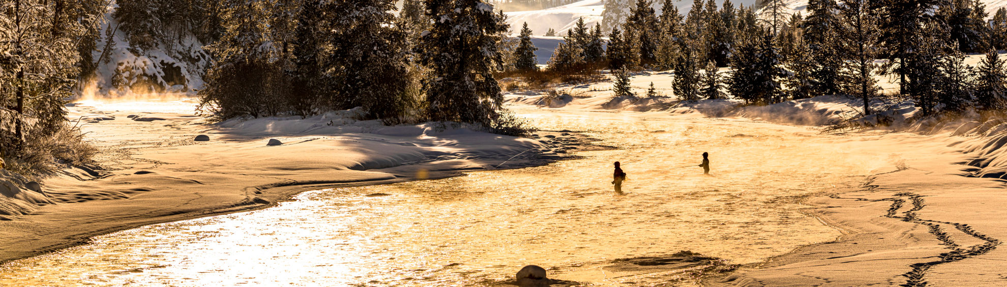 A stunning golden landscape at sunset with two anglers wading into a snowy stream.